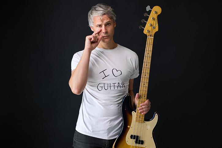 Gio holding a bass while wearing a t-shirt with the "I love guitar" inscription and looking sad