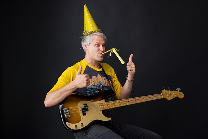 Picture of a celebratory Gio holding his bass, wearing a party hat and giving the thumbs up